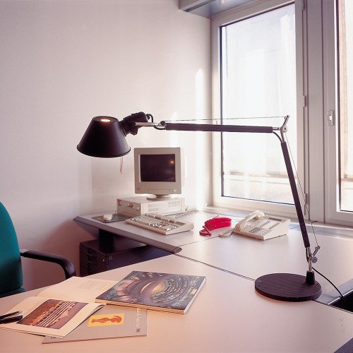 Tolomeo Mini negra trabajando sobre mesa de oficina junto a ordenador y documentos. Tolomeo Mini negra trabajando sobre mesa de oficina junto a ordenador y documentos.